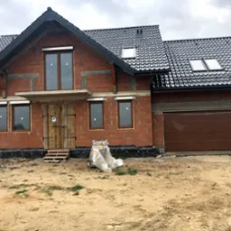 Surowy stan budynku jednorodzinnego z zamontowanymi brązowymi oknami i roletami zewnętrznymi, drewnianymi drzwiami wejściowymi i bramą garażową w kolorze brązowym, widoczne materiały budowlane...