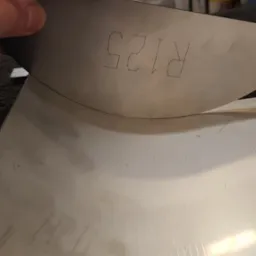 Metalowy element z wyciętym laserowo napisem 'R125', trzymany w dłoni, widoczne ślady obróbki na powierzchni.