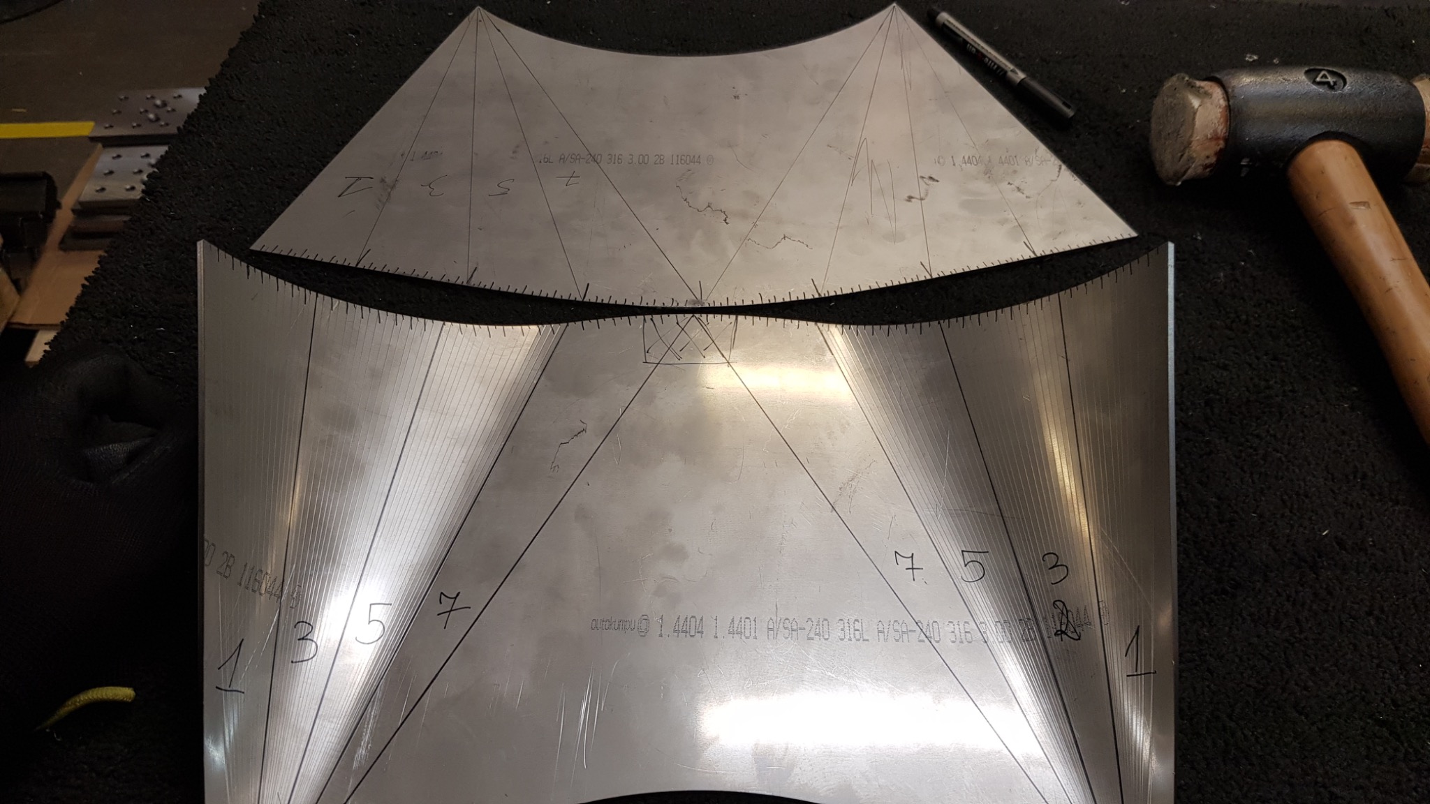 Dwie wygięte, metalowe płyty z naniesionymi liniami i numeracją, leżące na czarnym podłożu, obok młotek i długopis; prawdopodobnie etap przygotowania do obróbki metalu.