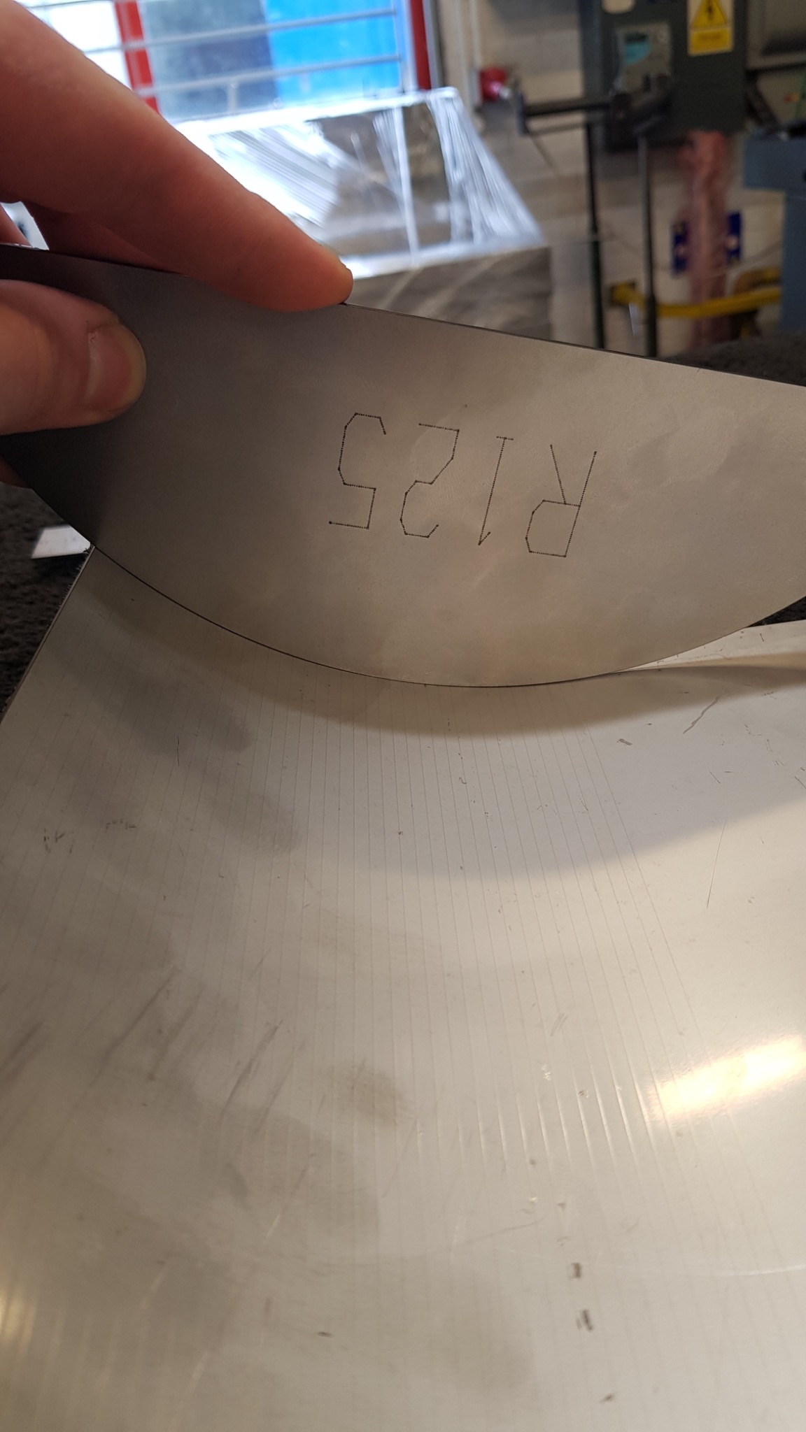 Metalowy element z wyciętym laserowo napisem 'R125', trzymany w dłoni, widoczne ślady obróbki na powierzchni.