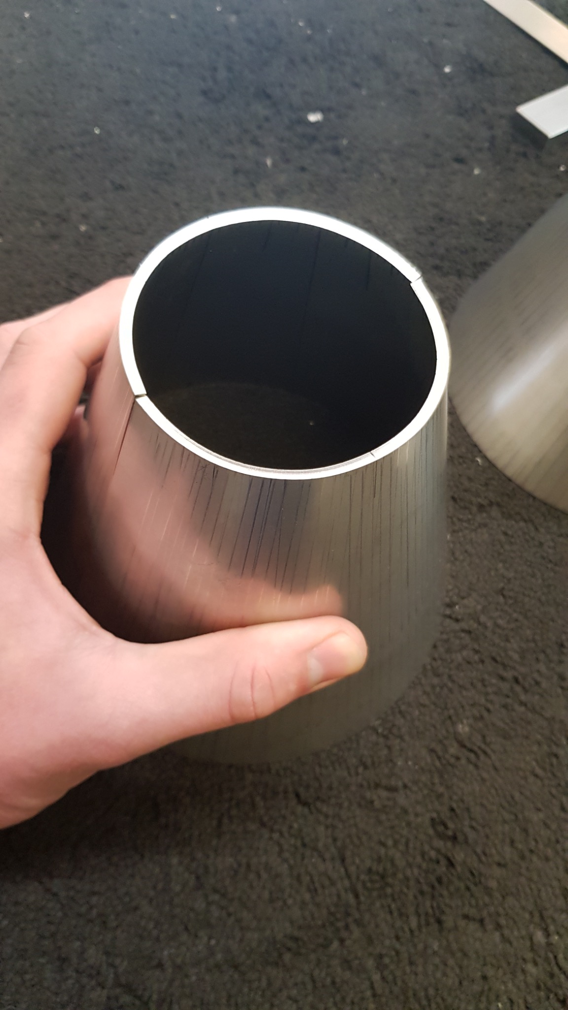 Obrabiany metalowy stożek w dłoni, widoczne ślady obróbki skrawaniem, drugi stożek w tle na szarym dywanie.