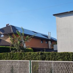 Firma Handlowo-Usługowa Patryk Szostak - Montaż paneli na dachu domu jednorodzinnego z drabiny; widoczne kominy i ogrodzenie.