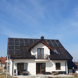 Firma Handlowo-Usługowa Patryk Szostak - Biały dom jednorodzinny z ciemną dachówką i panelami na dachu. Na podwórku stoi bus z otwartymi drzwiami i drabina oparta o ścianę.