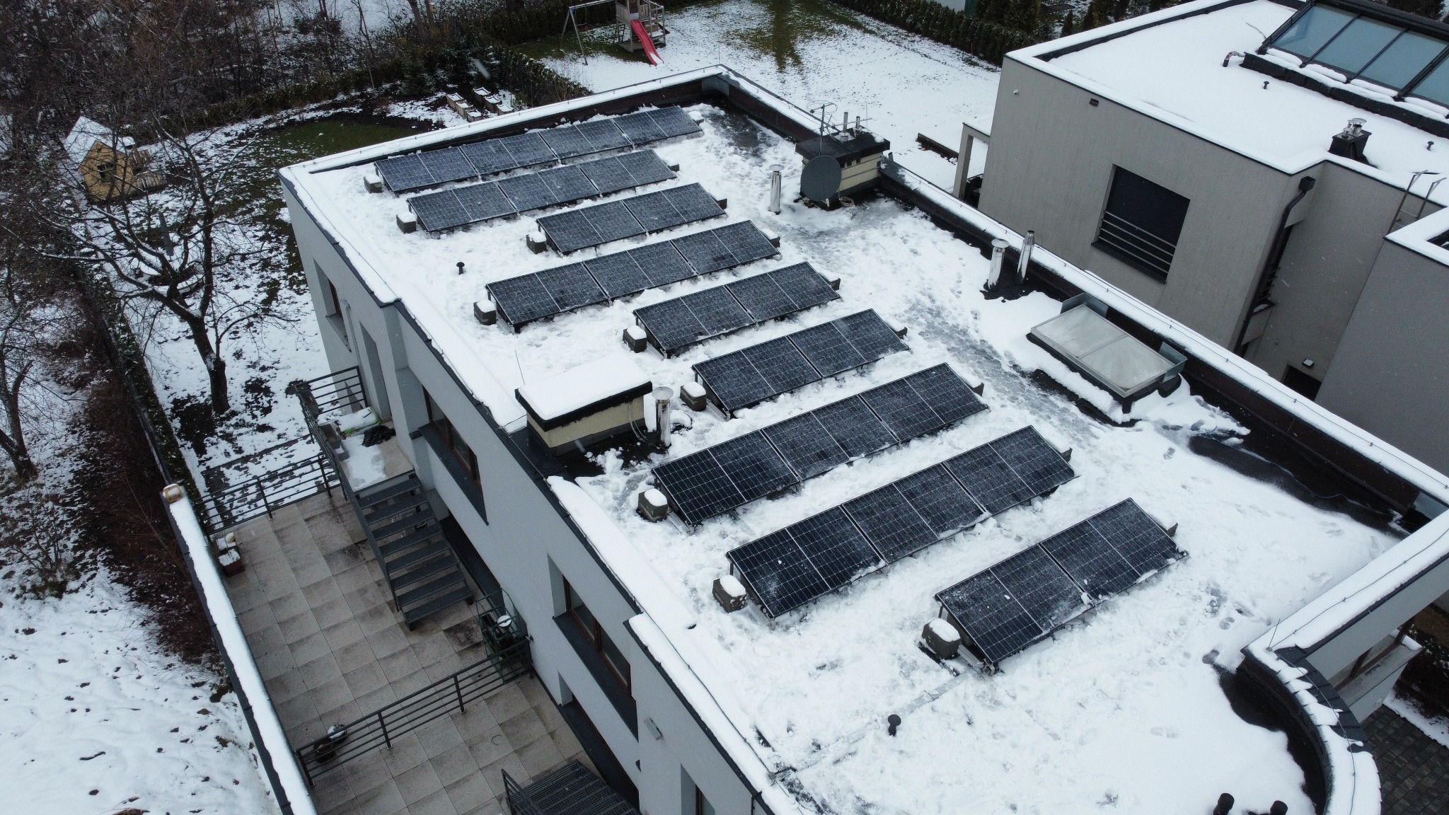 Zimowy widok z góry na panele słoneczne pokryte śniegiem na płaskim dachu nowoczesnego domu z jasną elewacją. Na dachu widoczne elementy instalacji i ślady butów w śniegu. W tle ośnieżony ogród...