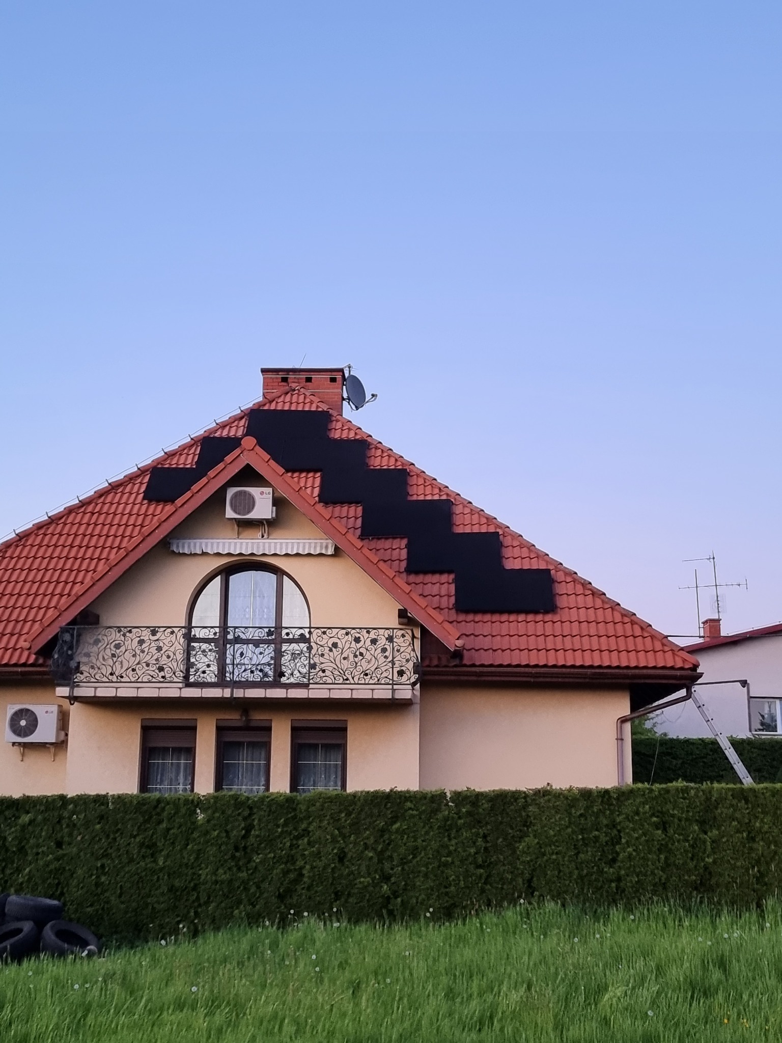 Instalacja paneli słonecznych na dachu domu jednorodzinnego z czerwoną dachówką, widok z poziomu trawnika, z klimatyzatorami na elewacji.