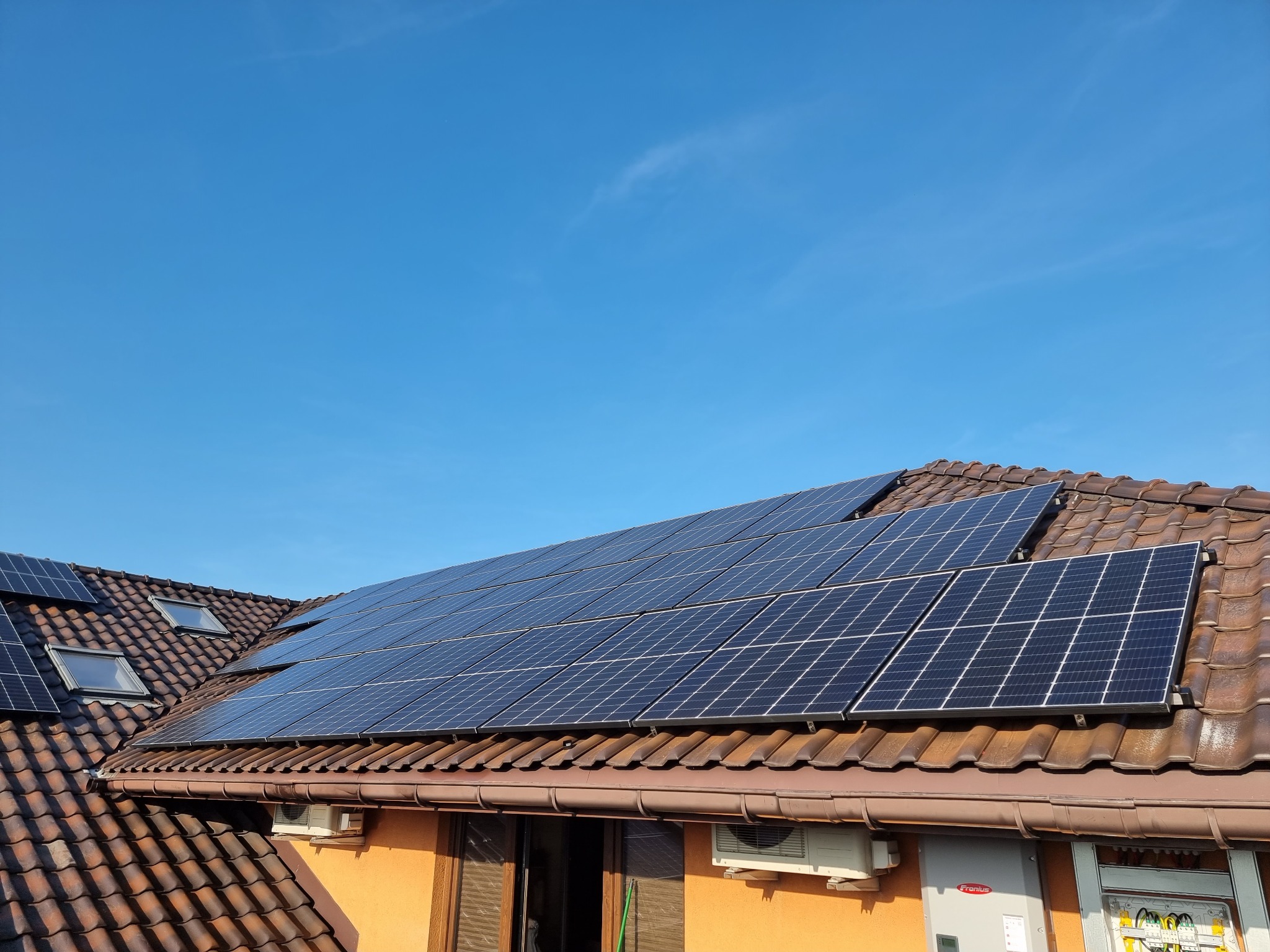 Panele fotowoltaiczne na brązowym dachu z dachówki ceramicznej, widoczne okna dachowe i klimatyzator na elewacji budynku, błękitne niebo w tle.