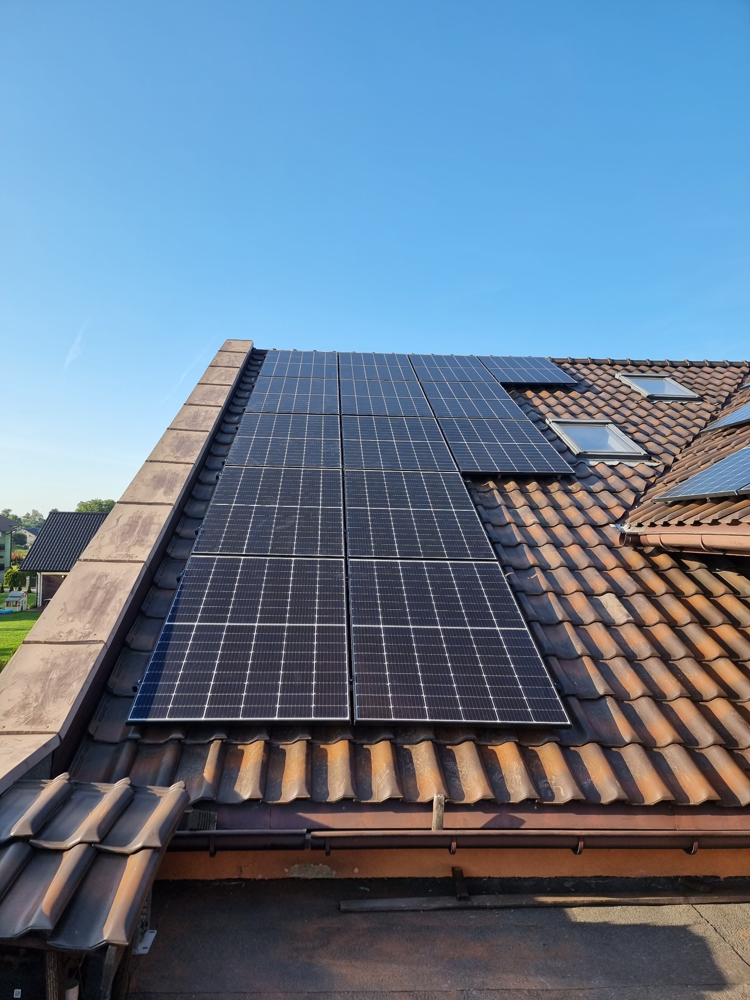 Panele słoneczne na dachu z brązowej dachówki, widoczne okna dachowe i fragmenty sąsiednich budynków na tle błękitnego nieba.