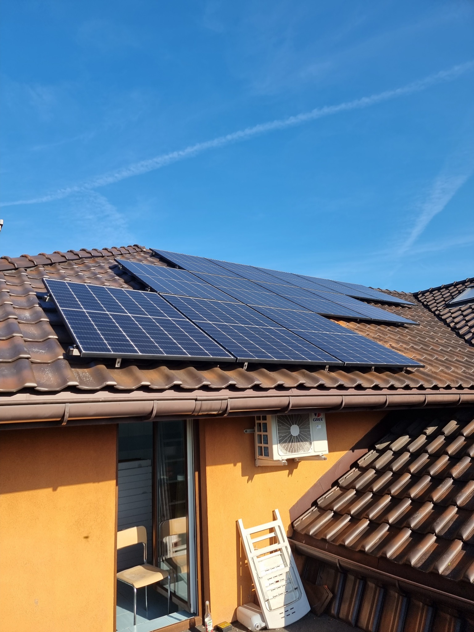 Panele słoneczne na brązowym, spadzistym dachu domu z pomarańczową elewacją, widoczne również klimatyzator, krzesło i drabina.