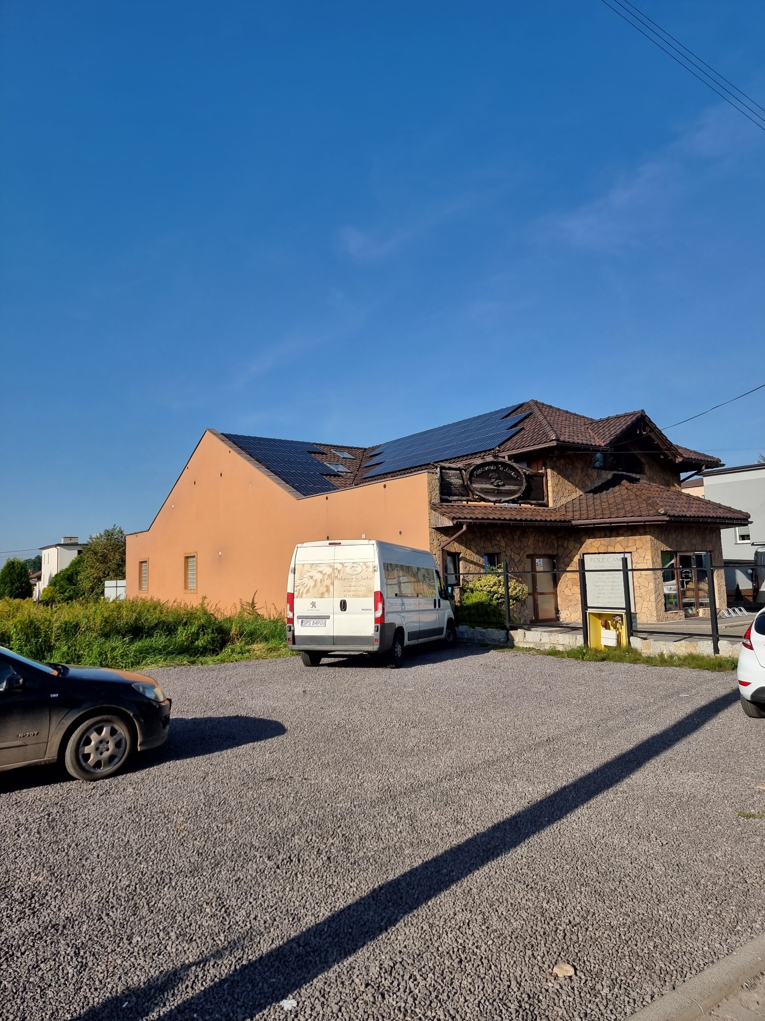 Instalacja paneli słonecznych na dachu budynku o nietypowej architekturze, z zaparkowanym białym dostawczakiem w tle, widok z poziomu żwirowego parkingu.