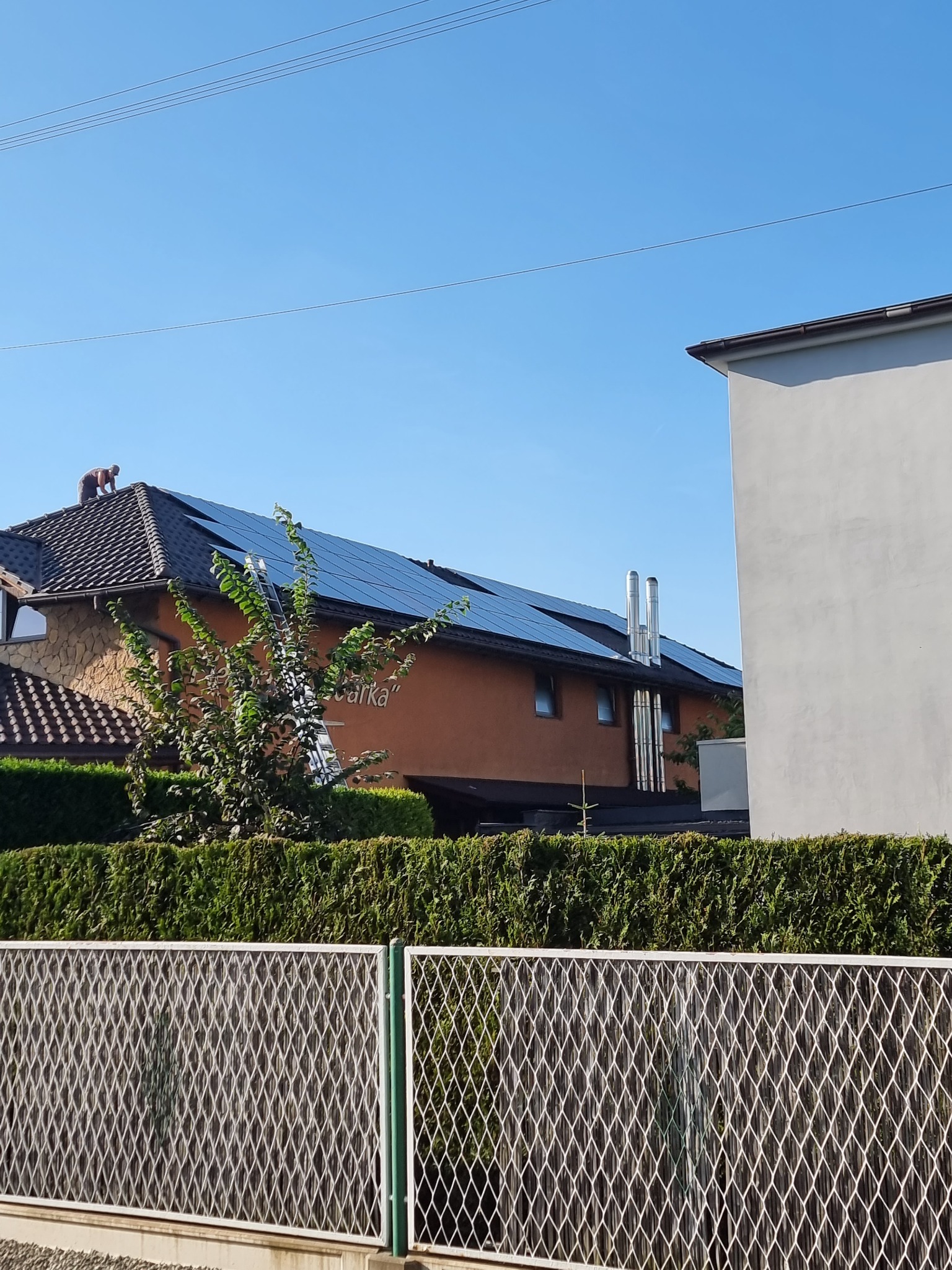 Montaż paneli na dachu domu jednorodzinnego z drabiny; widoczne kominy i ogrodzenie.