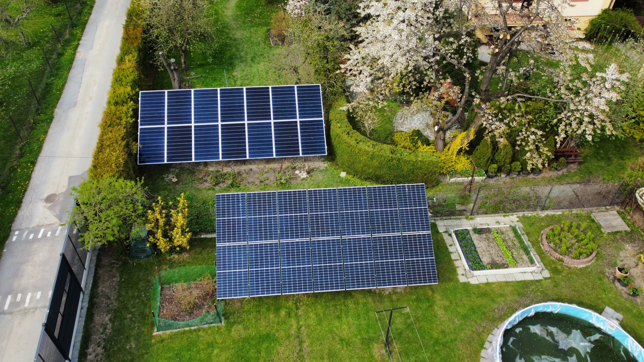 Dwa zestawy paneli słonecznych na trawniku w ogrodzie, widok z góry, obok drzewa kwitnącego na biało i mały basen.