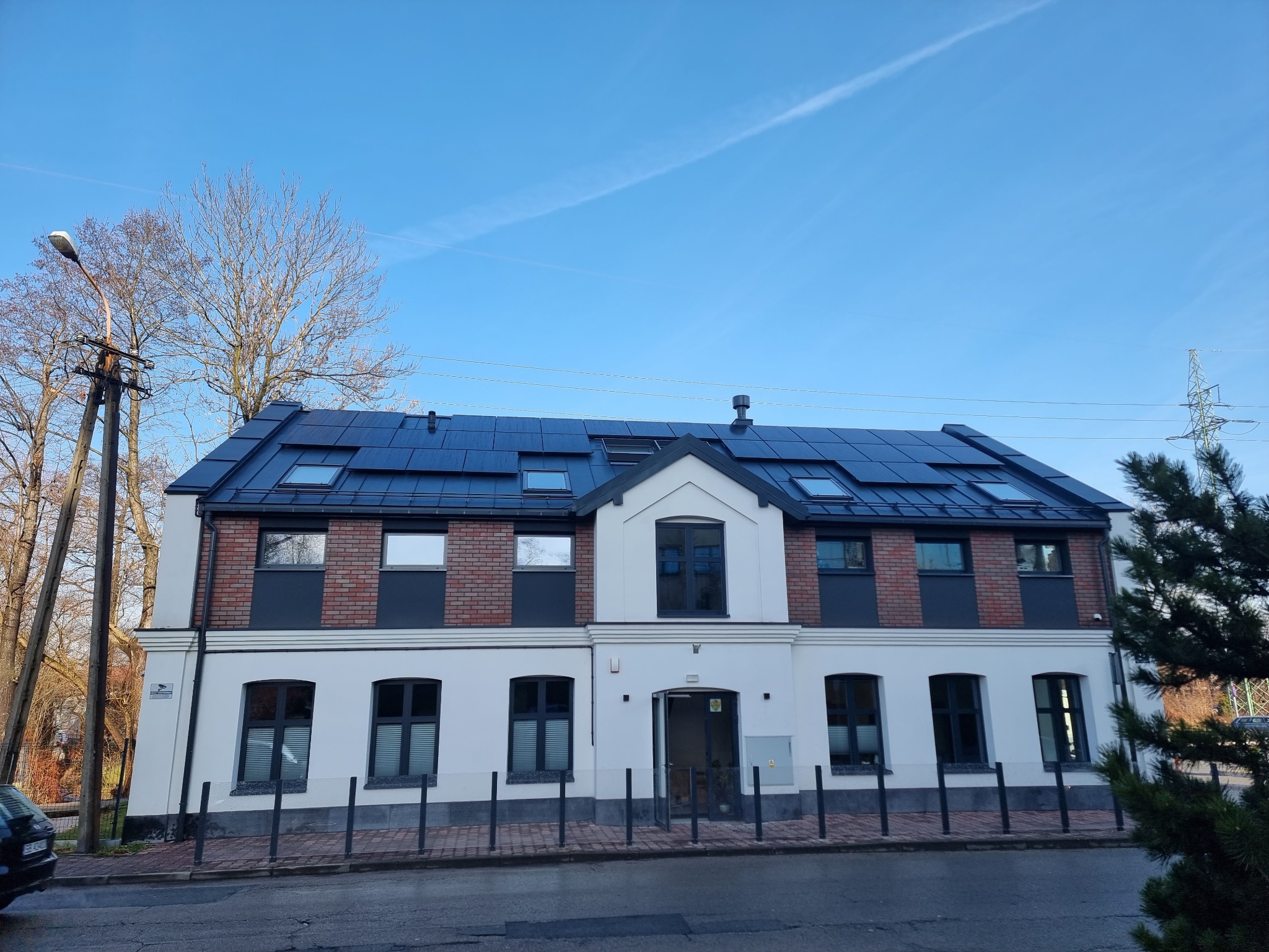 Dwupiętrowy budynek z ciemnymi panelami na dachu, oświetlony słonecznym światłem, z widocznym fragmentem nieba i drzew