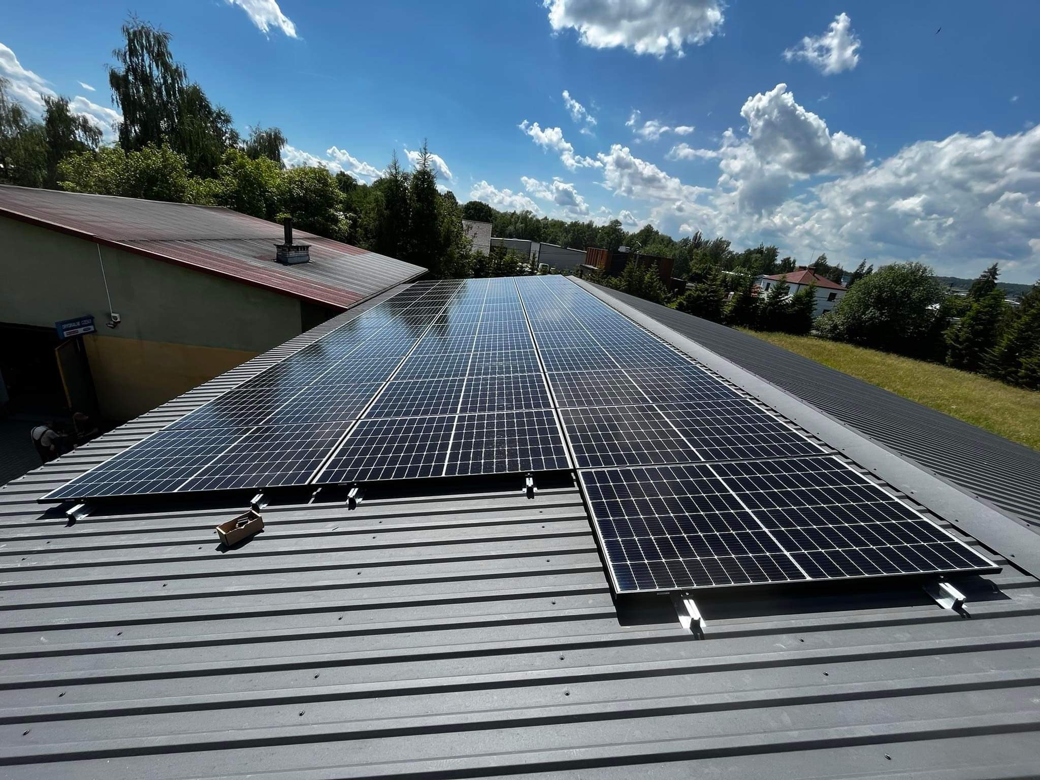 Panele fotowoltaiczne na szarym, metalowym dachu z widocznymi elementami montażowymi, na tle błękitnego nieba z białymi chmurami i zieleni drzew. Na dachu leży kartonowe pudełko.