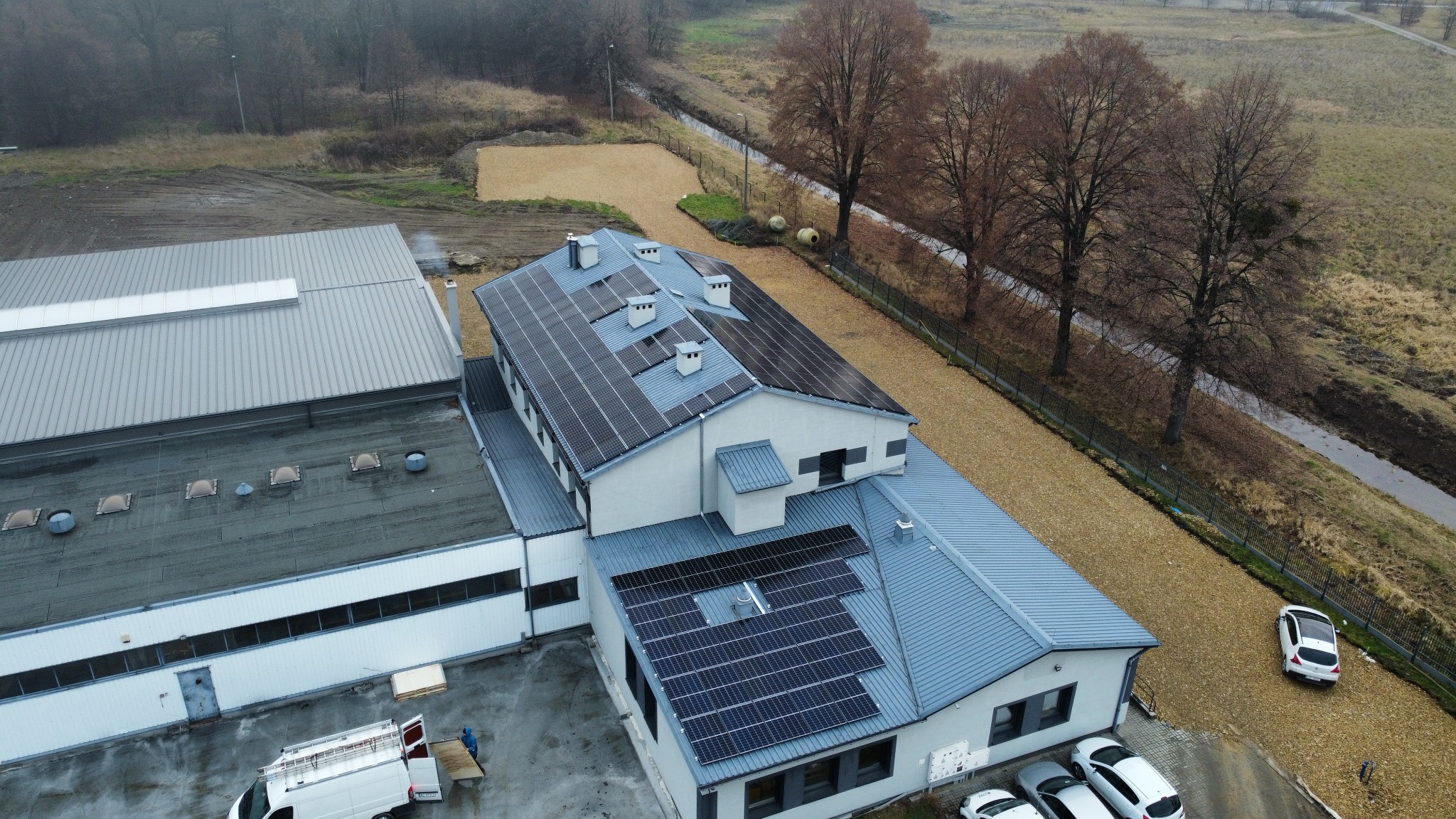Widok z drona na budynek z panelami słonecznymi na dachu, otoczony żwirowym terenem i drzewami bez liści, z zaparkowanymi samochodami i furgonetką w pobliżu.