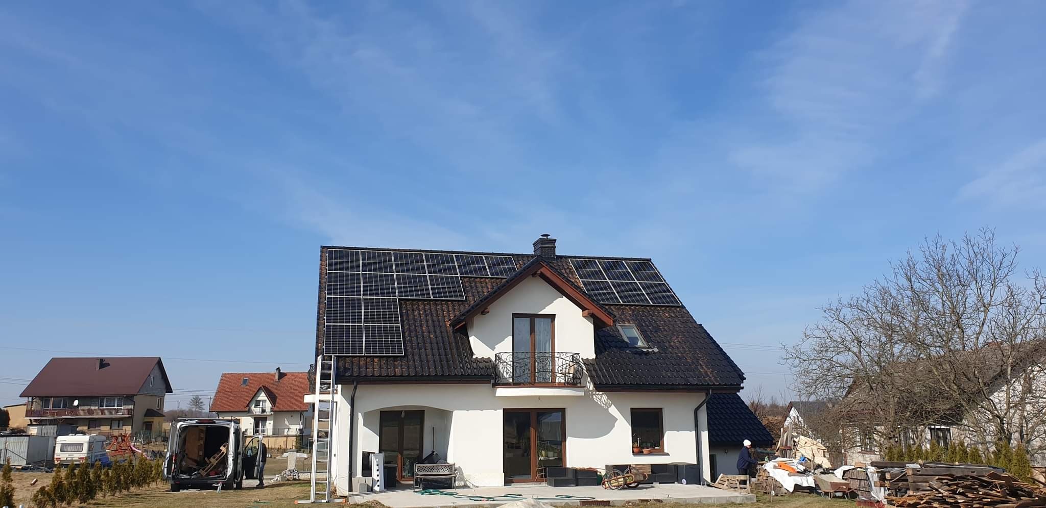 Biały dom jednorodzinny z ciemną dachówką i panelami na dachu. Na podwórku stoi bus z otwartymi drzwiami i drabina oparta o ścianę.