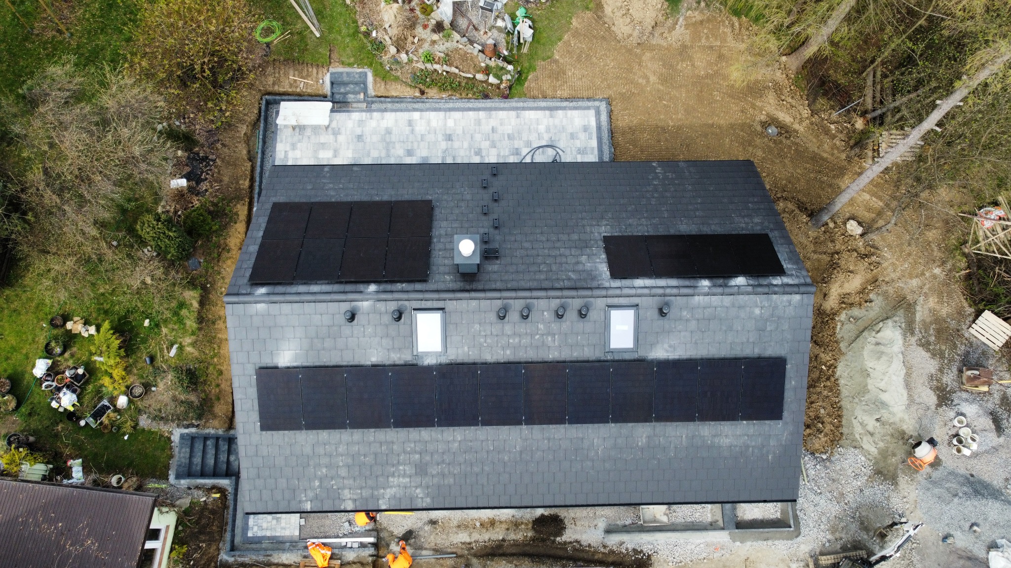 Widok z drona na dach domu z panelami słonecznymi, dwoma oknami dachowymi i kominem wentylacyjnym. Część paneli już zamontowana, a obok dachu widoczni pracownicy w pomarańczowych kamizelkach. Teren...