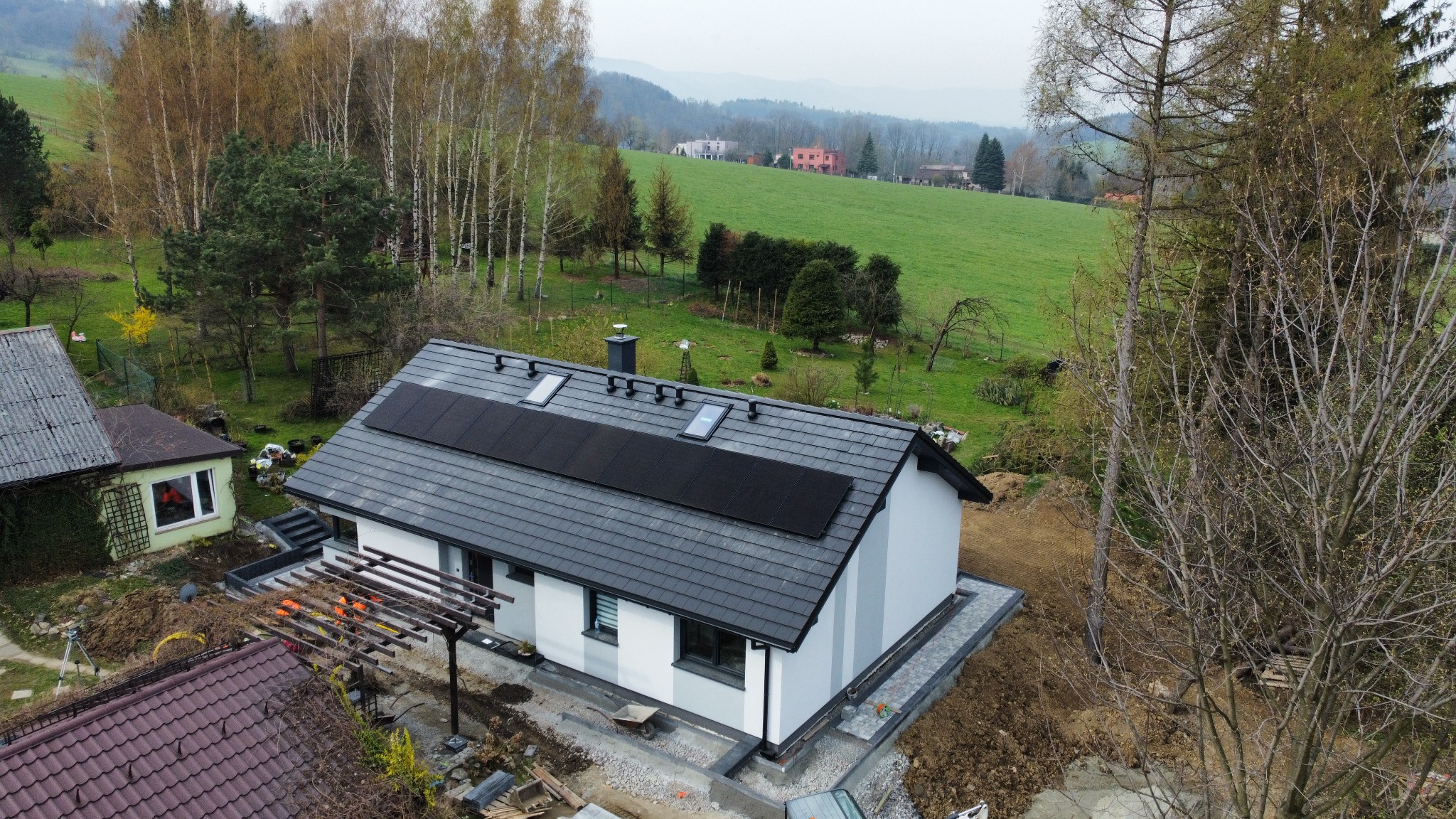Nowoczesny dom jednorodzinny z szarym dachem i panelami słonecznymi, widok z góry na tle zielonego krajobrazu. Trwają prace wykończeniowe wokół budynku.