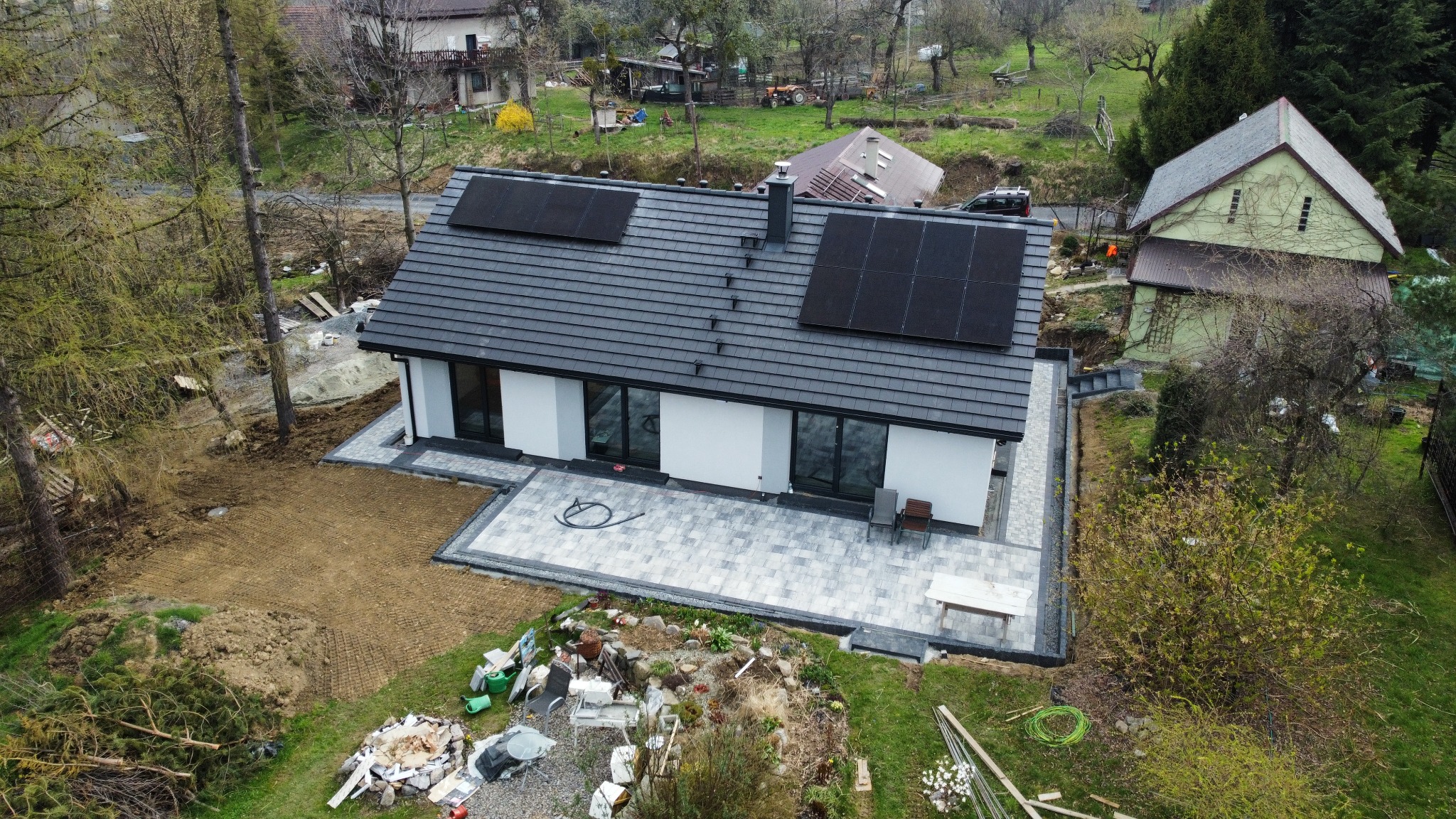Nowoczesny dom jednorodzinny z panelami fotowoltaicznymi na dachu, widok z góry, świeżo ułożony taras z kostki brukowej, teren wokół domu w trakcie zagospodarowania.