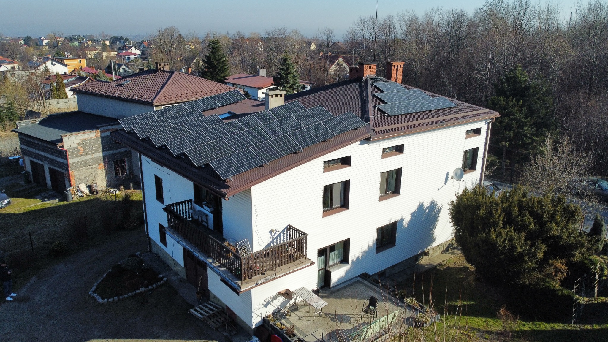 Widok z góry na dom jednorodzinny z panelami słonecznymi na brązowym dachu, otoczony drzewami i zabudową osiedlową.