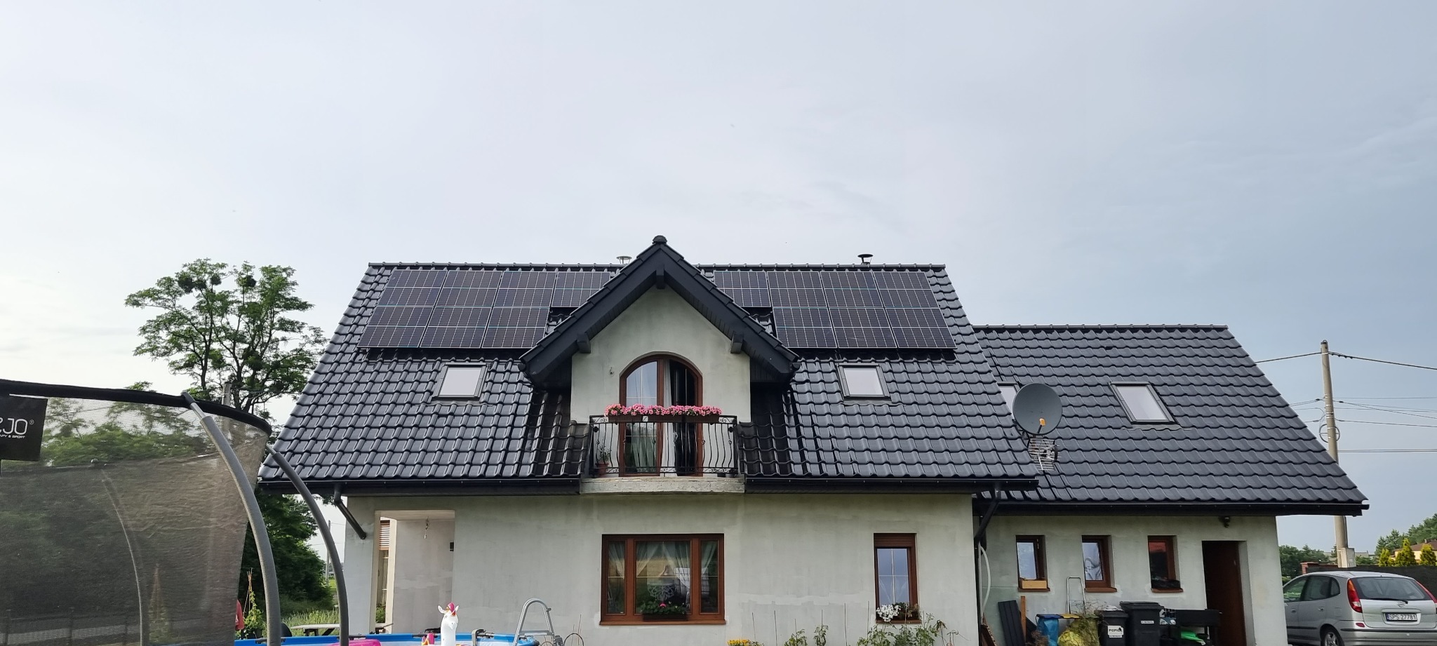 Dom jednorodzinny z ciemną dachówką i panelami słonecznymi na dachu, widok z poziomu terenu, w tle pochmurne niebo.