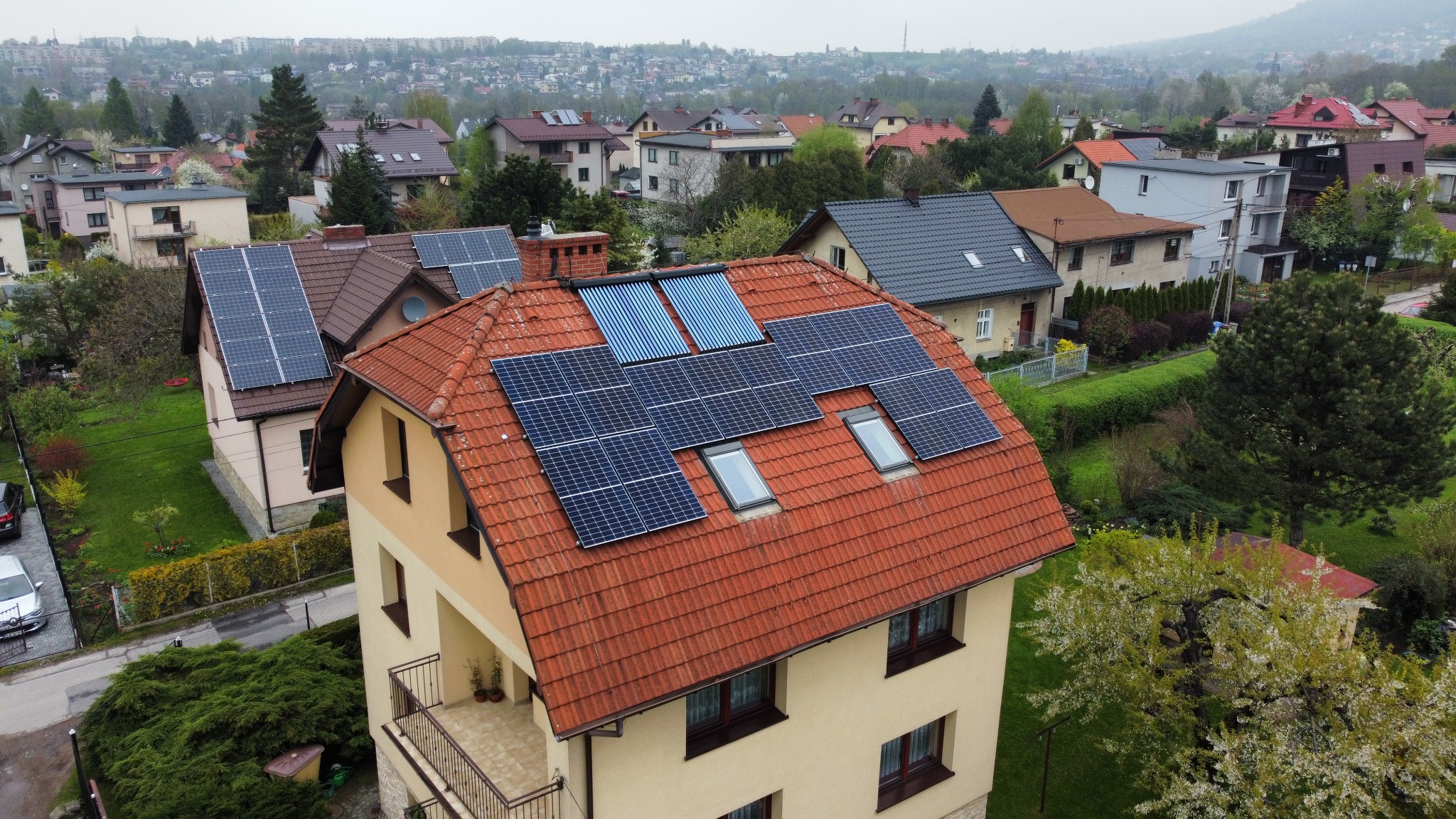 Dom jednorodzinny z czerwoną dachówką, na której zamontowano panele fotowoltaiczne i kolektory słoneczne, widok z góry na tle zabudowy miejskiej i wzgórz.