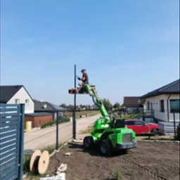 Pracownik na podnośniku montuje słupek ogrodzeniowy z instalacją elektryczną, widoczne bębny z kablem, domy w tle.