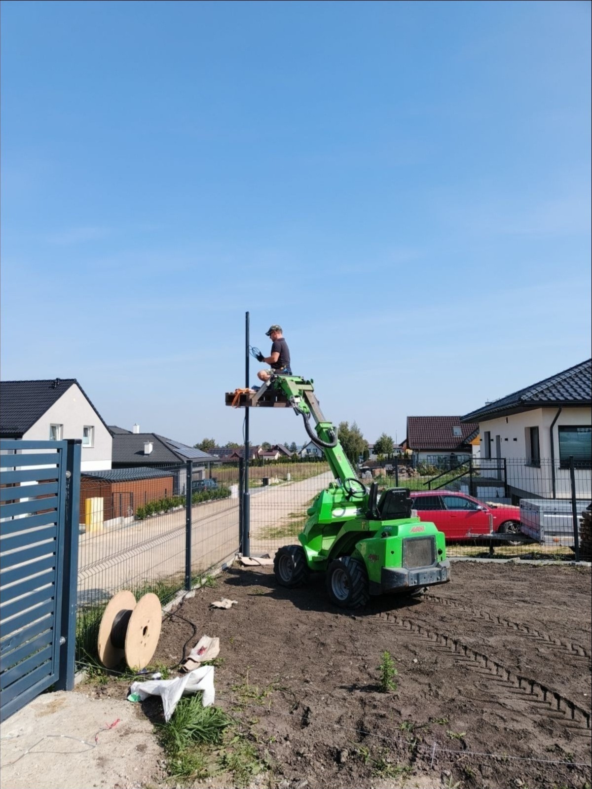 Pracownik na podnośniku montuje słupek ogrodzeniowy z instalacją elektryczną, widoczne bębny z kablem, domy w tle.