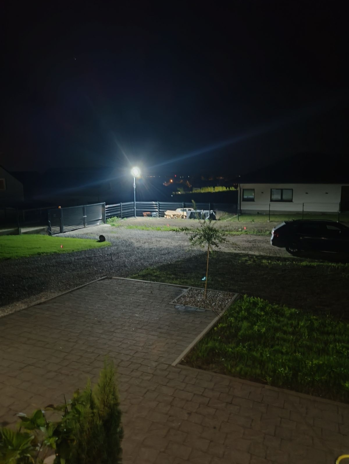 Oświetlony teren posesji nocą, widoczna lampa uliczna, trawnik, podjazd z kostki brukowej, ogrodzenie, budynek mieszkalny i zaparkowany samochód.