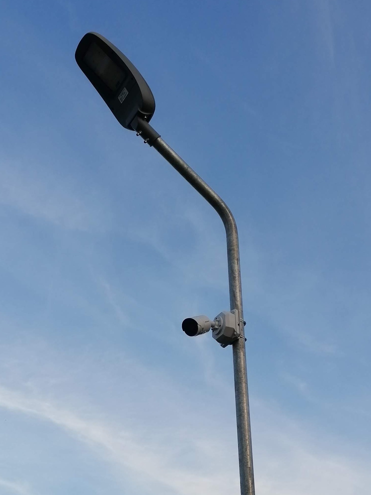 Słup oświetleniowy z zamontowaną kamerą przemysłową na tle błękitnego nieba z delikatnymi obłokami.