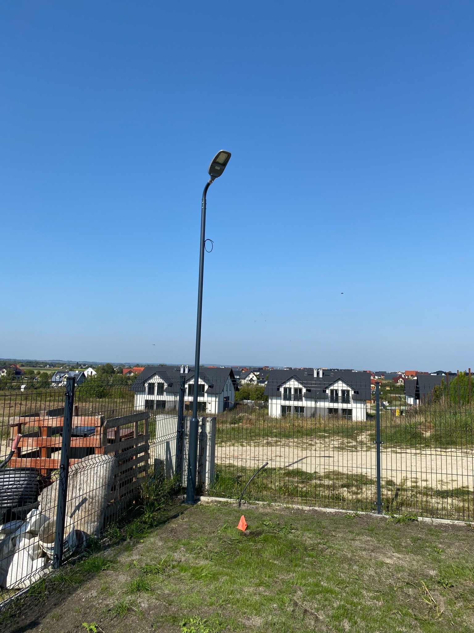 Słup oświetleniowy na tle nowo wybudowanych domów jednorodzinnych, widoczny zza ogrodzenia z siatki.