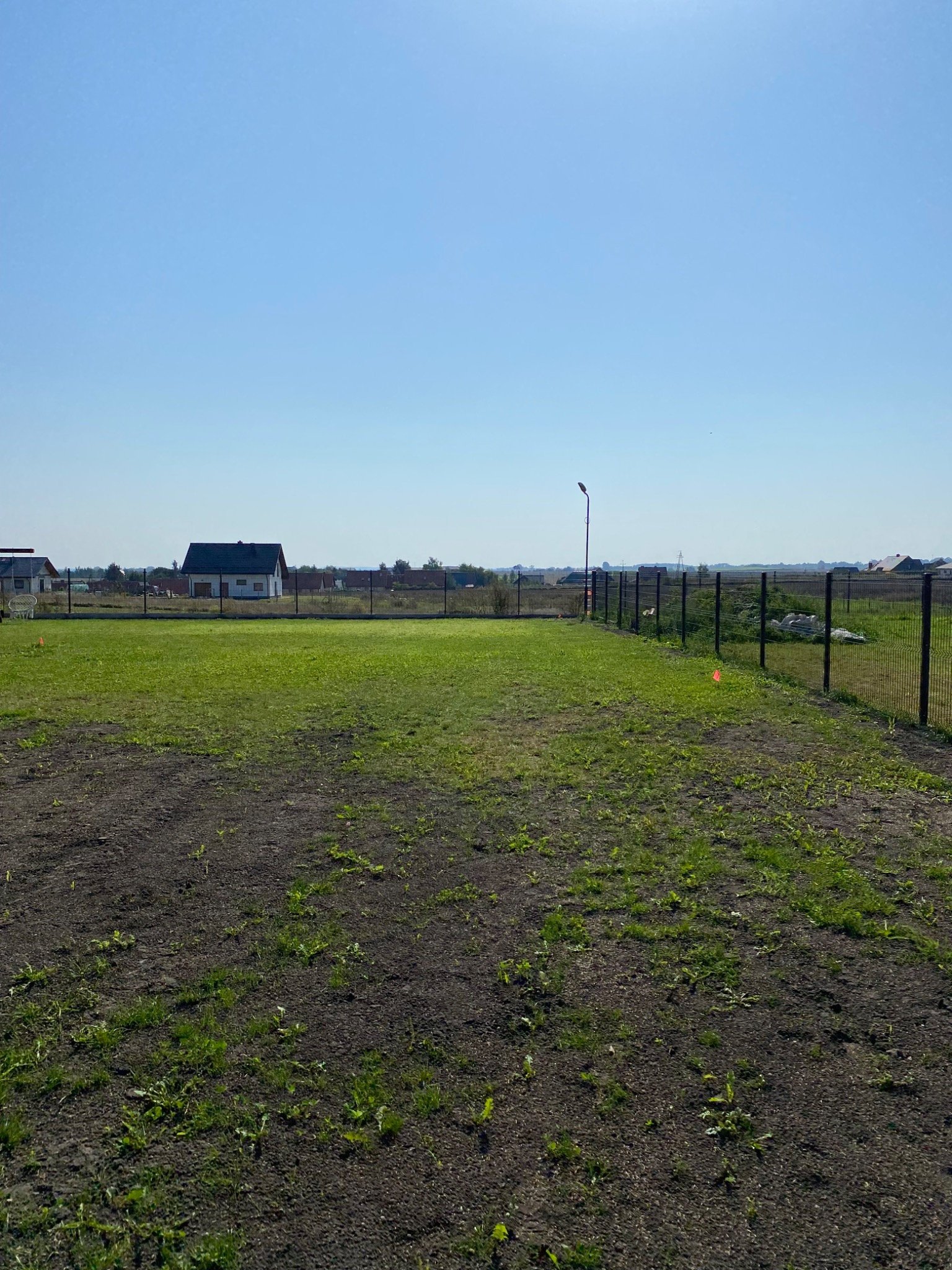 Działka z nowo postawionym, ciemnym ogrodzeniem panelowym na podmurówce, widok na domy w oddali, słoneczny dzień.