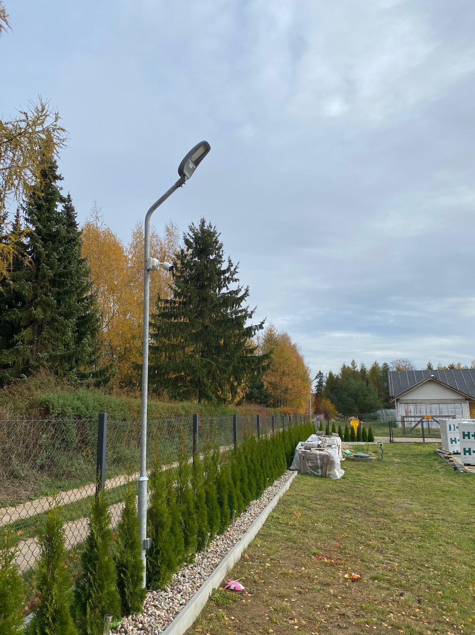 Słup oświetleniowy z zamontowaną kamerą na posesji, widok na ogrodzenie z tujami i dom w tle.