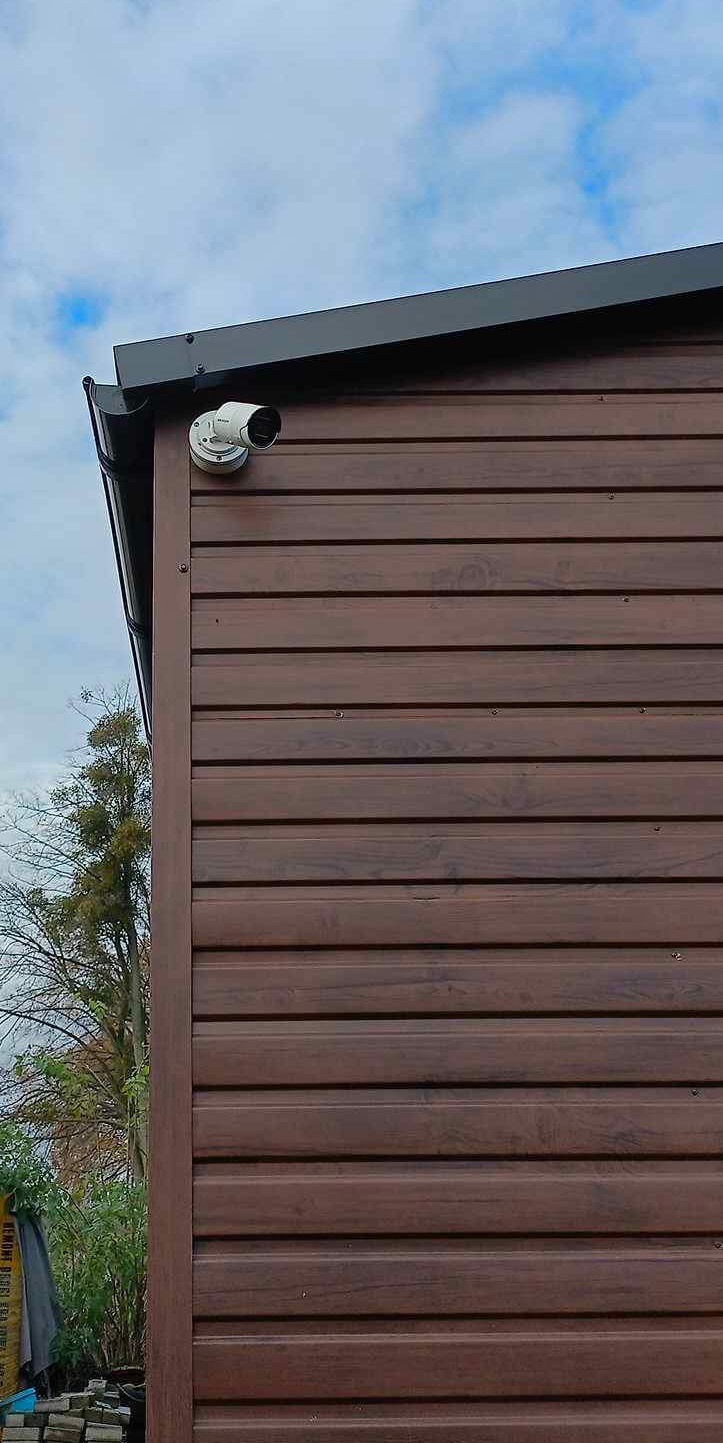 Kamera monitoringu zamontowana na brązowej elewacji budynku z drewnianą fakturą, widok z dołu na tle błękitnego nieba.