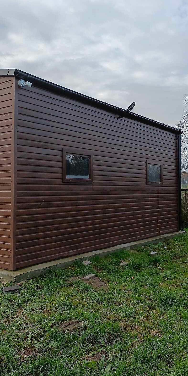 Brązowy budynek gospodarczy z dwoma małymi oknami i zamontowaną kamerą na rogu, otoczony trawą.