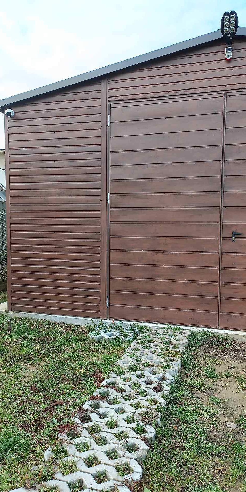 Brązowy garaż blaszany z oświetleniem LED i kamerą monitoringu, do którego prowadzi ścieżka z ażurowych betonowych płyt.