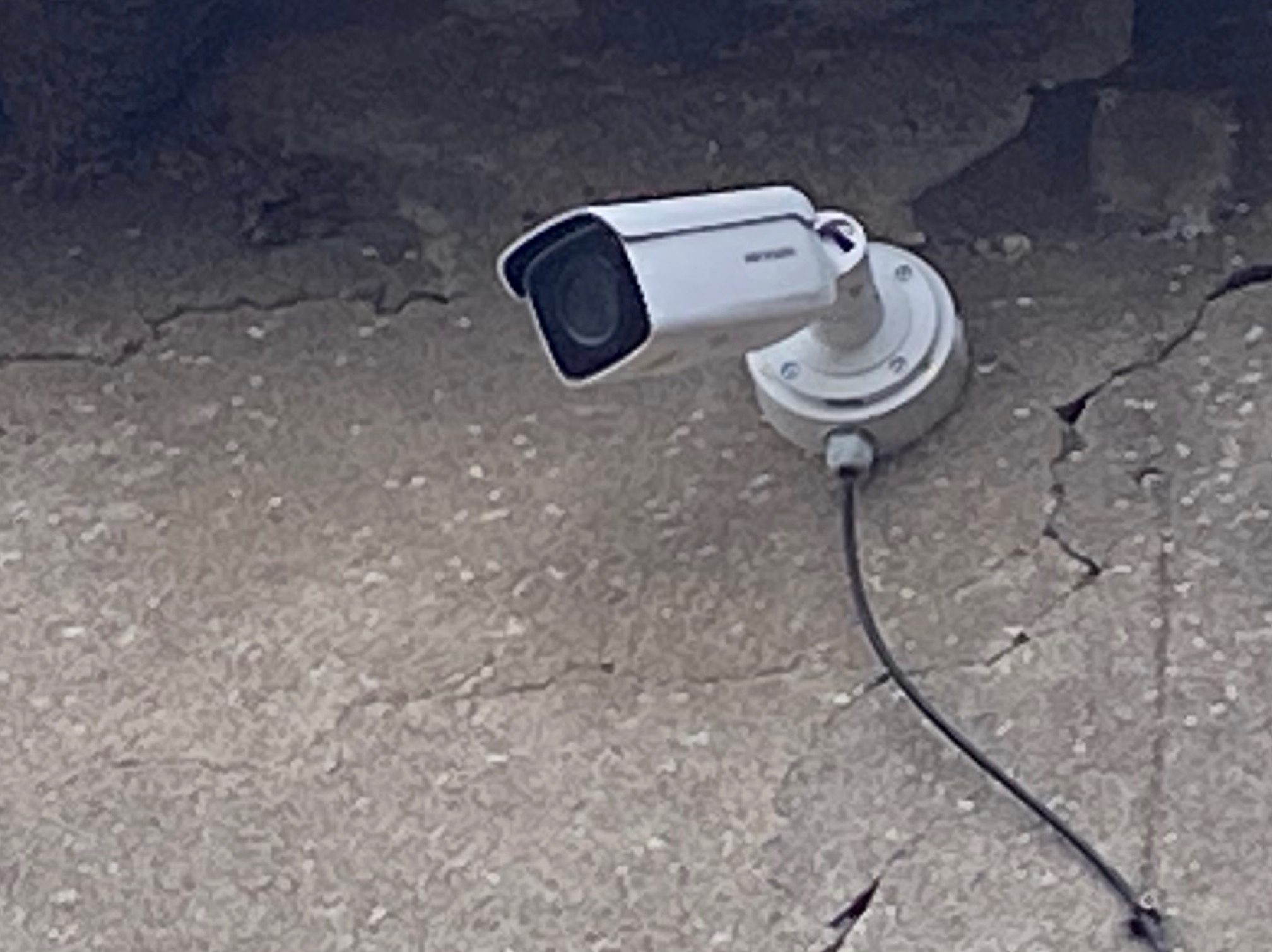 Biała kamera monitoringu zamontowana na szarej, popękanej ścianie zewnętrznej budynku. Widoczny kabel zasilający.