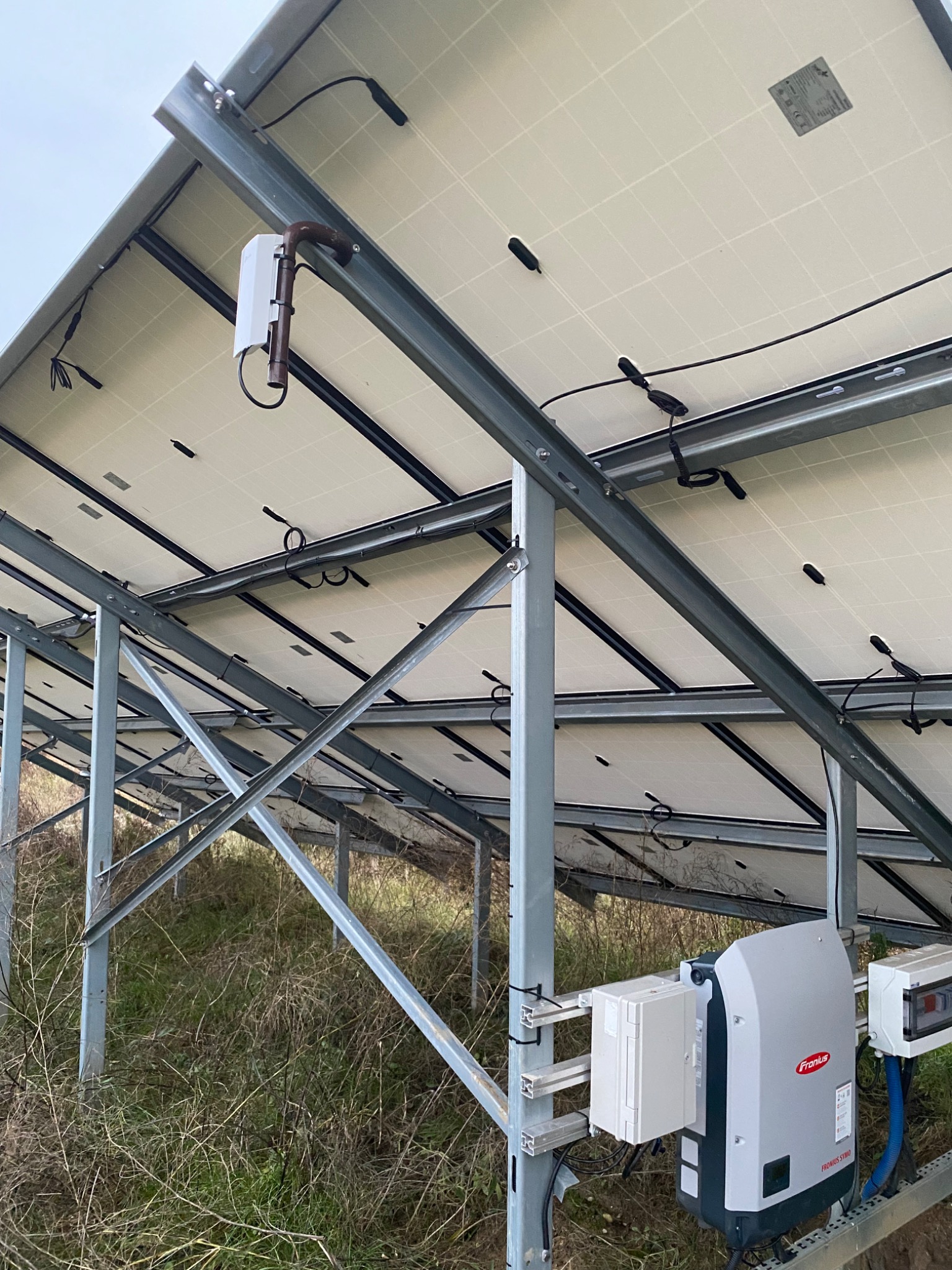 Widok od spodu na panele fotowoltaiczne zamontowane na metalowej konstrukcji, widoczny inwerter firmy Fronius oraz okablowanie.