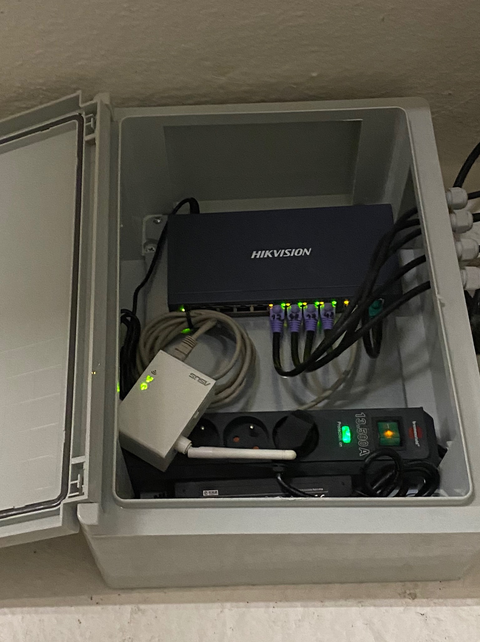 Switch sieciowy Hikvision i router ASUS w skrzynce instalacyjnej z listwą zasilającą Brennenstuhl, widoczne kable i diody sygnalizacyjne.