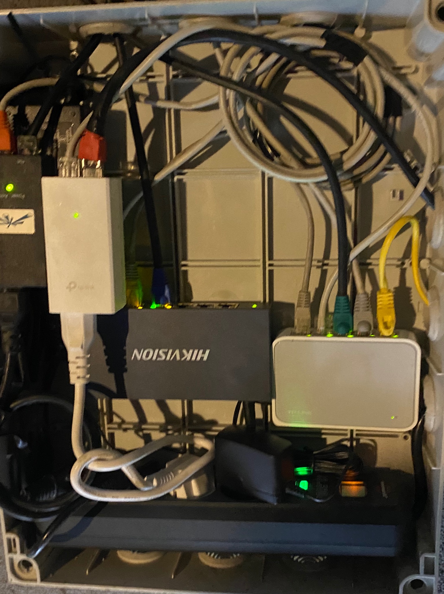 Rozdzielnica z widocznymi kablami sieciowymi, zasilaczami i elementami elektroniki, w tym router Hikvision z podłączonymi kablami Ethernet różnych kolorów, oraz zasilacz TP-Link z diodami LED...