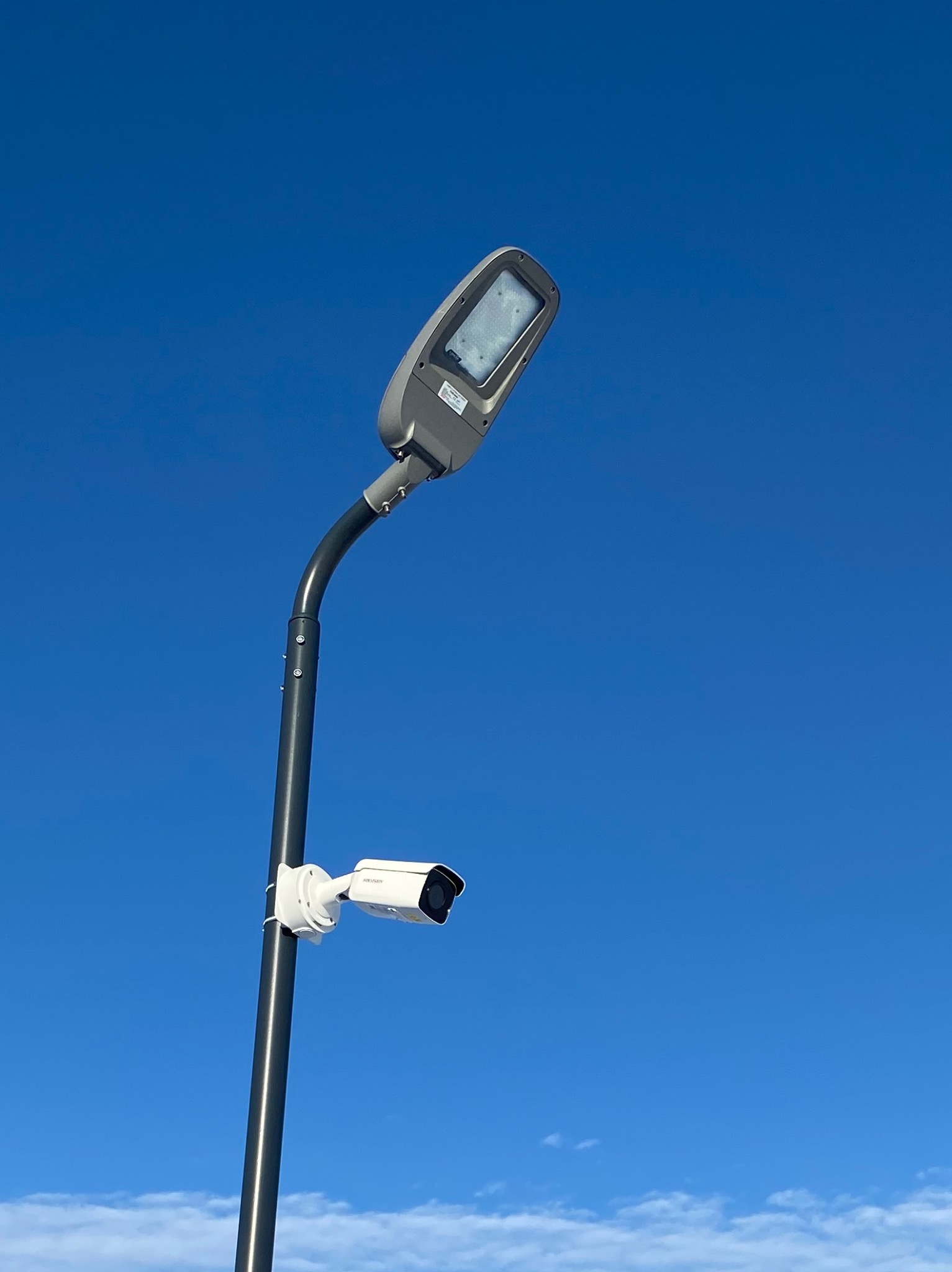 Lampa uliczna LED z zamontowaną kamerą monitoringu na tle błękitnego nieba z delikatnymi obłokami.