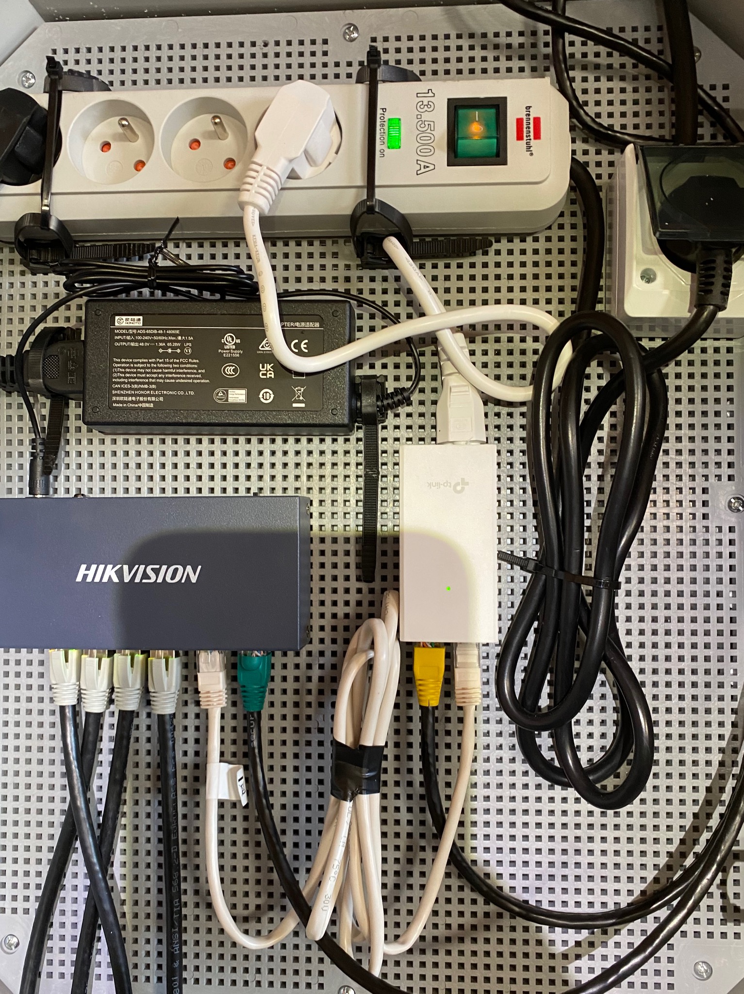 Panel krosowniczy z urządzeniami sieciowymi Hikvision i TP-Link, zasilaczem, listwą zasilającą Brennenstuhl oraz okablowaniem Ethernet, wszystko zamontowane na perforowanej płycie montażowej.