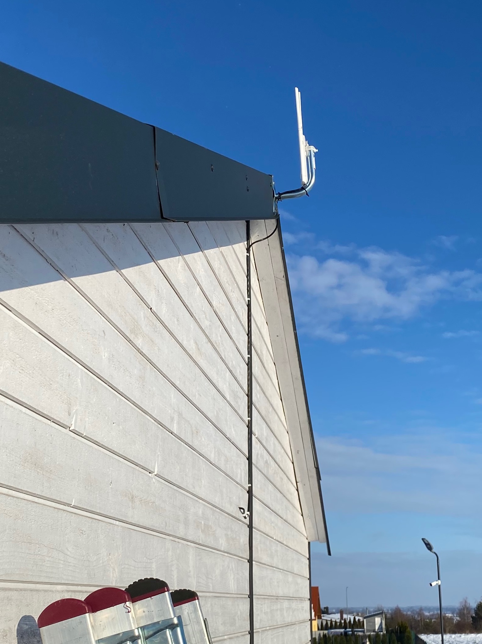 Zewnętrzna antena na elewacji domu z białego sidingu, zamontowana na metalowym wsporniku, z widocznymi kablami prowadzonymi wzdłuż ściany, drabina oparta o ścianę, błękitne niebo w tle.