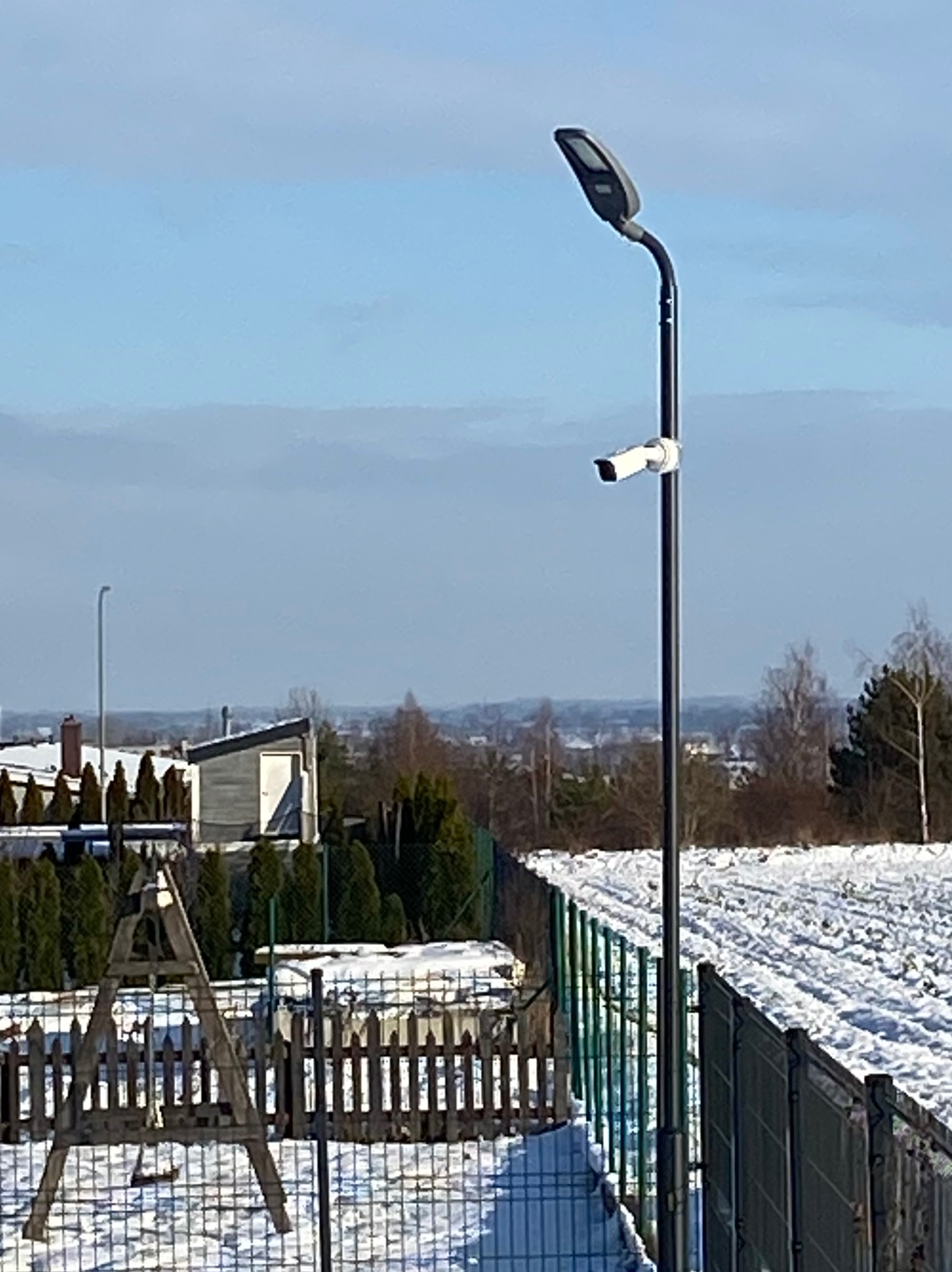 Kamera monitoringu zamontowana na słupie oświetleniowym na tle zimowego krajobrazu z ogrodzeniem i zabudowaniami.
