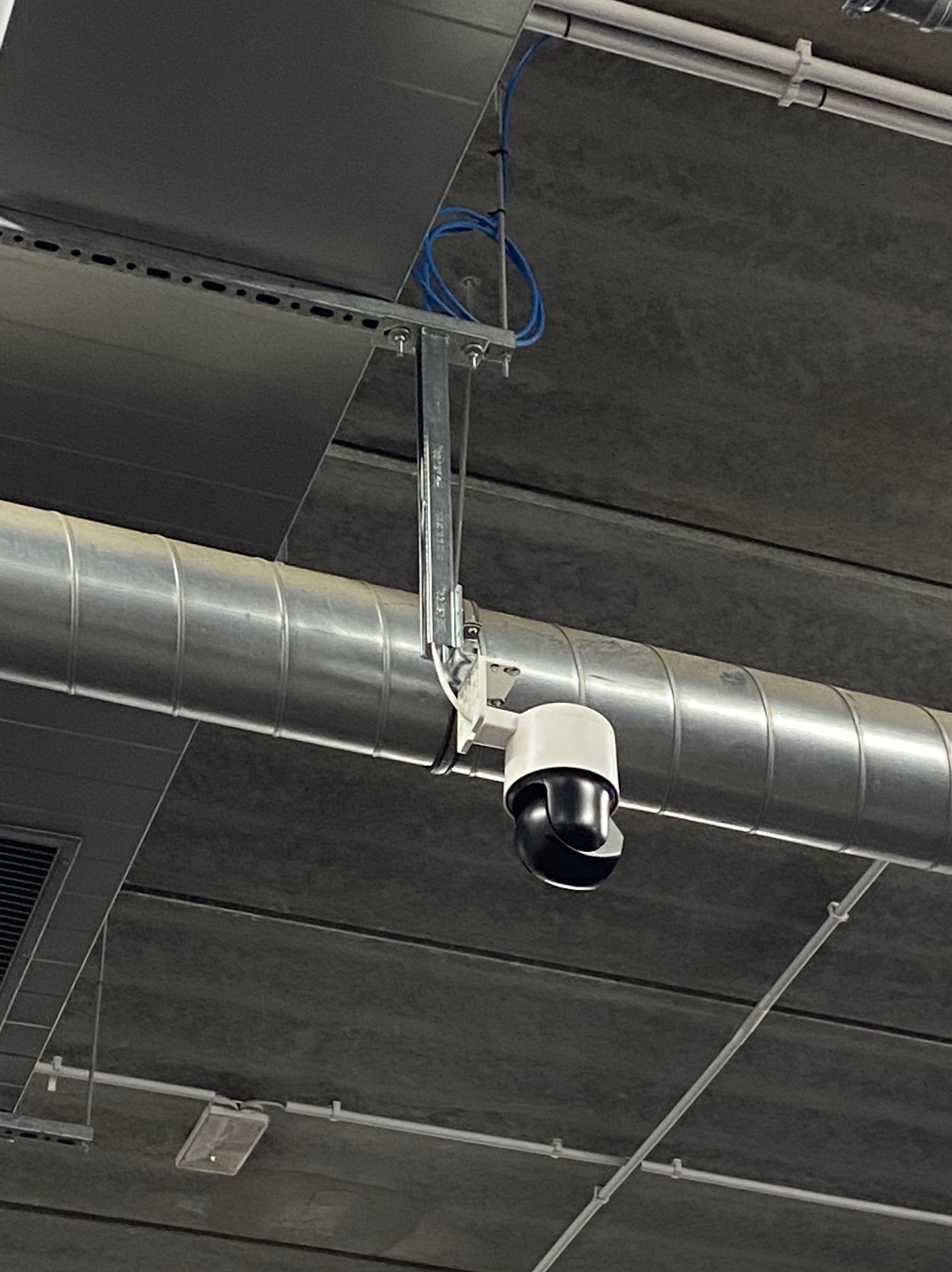 Kamera przemysłowa PTZ zamontowana na metalowej rurze wentylacyjnej pod sufitem, widoczne niebieskie kable i elementy mocujące.