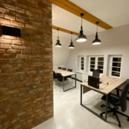 Wykończone biuro z ceglaną ścianą, drewnianymi belkami, wiszącymi lampami, jasnymi ścianami i podłogą oraz nowoczesnymi biurkami z komputerami.