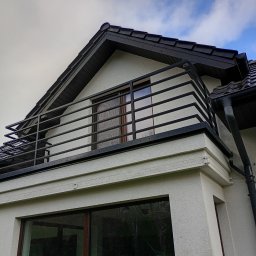 Andrii Dets - Fragment elewacji domu z balkonem z czarną, metalową balustradą i oknem na piętrze, widok z dołu na tle pochmurnego nieba.