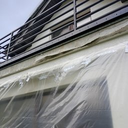Andrii Dets - Fragment elewacji domu z balkonem w trakcie prac, zabezpieczony folią ochronną, widoczna siatka zbrojąca.