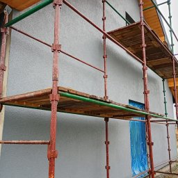 Andrii Dets - Budynek jednorodzinny w trakcie prac elewacyjnych, widoczne rusztowanie z drewnianymi podestami, okno zabezpieczone folią, elewacja pokryta szarym tynkiem strukturalnym.