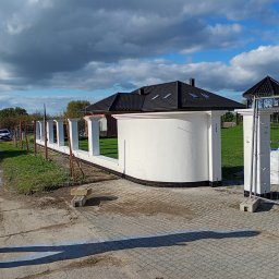 Andrii Dets - Nowo wybudowane, białe ogrodzenie z filarami i podmurówką z czarną glazurą, widoczne na tle domu z czarnym dachem i błękitnego nieba z chmurami.