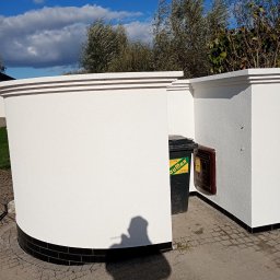 Andrii Dets - Biała, teksturowana ściana zewnętrzna z dekoracyjnymi listwami i czarną, ceramiczną bazą, osłaniająca pojemnik na śmieci i skrzynkę gazową, na tle błękitnego nieba z chmurami.